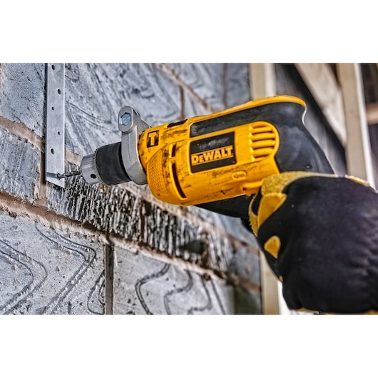 DEWALT drill in use, drilling into a metal bracket on a brick wall. Gloved hand holds the yellow power tool.