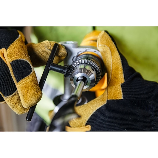 Gloved hands adjust a DEWALT drill with a chuck key for a drill bit. Close-up of tool and safety gloves.