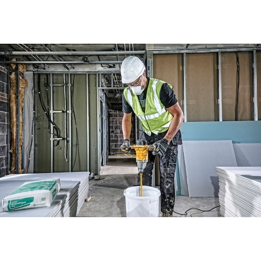 Worker uses a DEWALT DWD241 drill to mix materials in a bucket on a construction site.