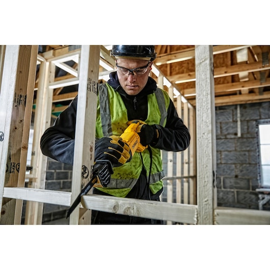 Worker uses a yellow DEWALT reciprocating saw to cut wood frame at a construction site, wearing safety gear.