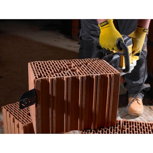 A person wearing yellow gloves is using a DEWALT power tool to cut through a large hollow clay brick. The tool is positioned against the brick and the person is standing on a dusty surface.