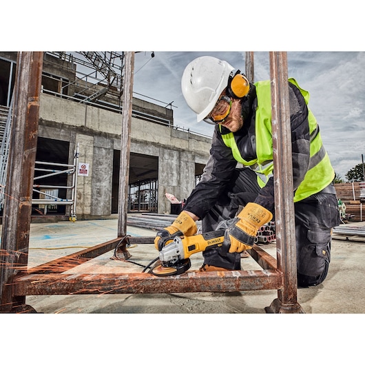 Worker uses DEWALT DWE4357 grinder on metal frame at construction site, wearing safety gear.