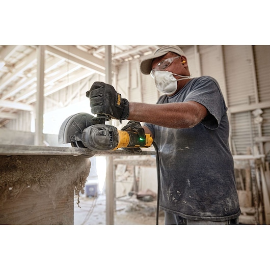 A worker wearing protective gear, including gloves, a mask, goggles, and earplugs, is using a DEWALT angle grinder to cut a piece of material in an industrial workshop setting.