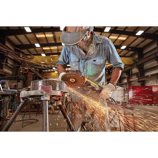 A worker wearing safety gear uses a DEWALT angle grinder, model DWE4377, to cut metal in an industrial workshop. Bright sparks are flying from the cutting area.