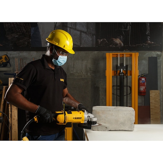 Person using a DEWALT grinder to cut concrete block in a workshop, wearing safety gear and gloves.