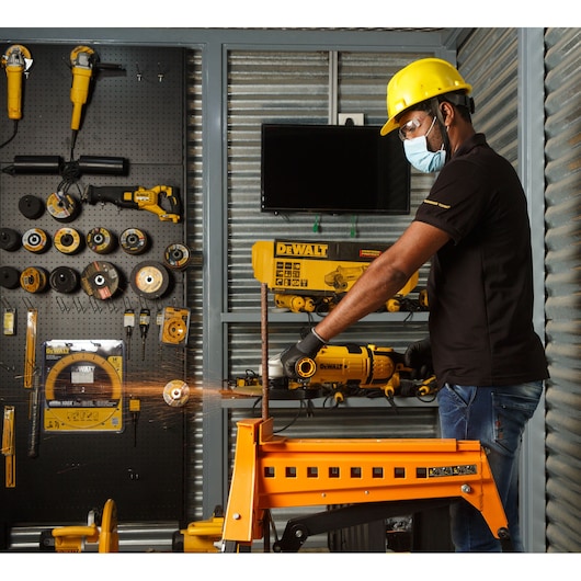 Worker uses DEWALT tool to cut metal in workshop, wearing safety gear. Displayed tools and equipment in background.