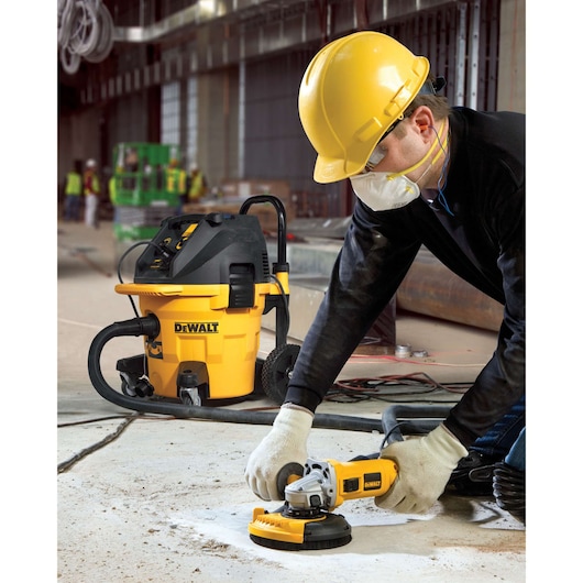 A construction worker wearing a yellow hard hat, gloves, and face mask is using a DEWALT DWE46150 surface grinding tool on a concrete floor, with a yellow DEWALT dust extractor vacuum in the background. The scene is set in an industrial or construction environment.