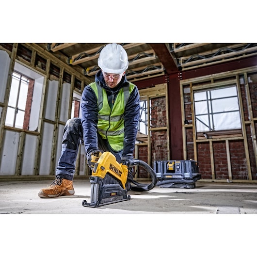 Worker uses DEWALT cutting tool on floor at construction site, wearing safety gear. Toolbox in background.