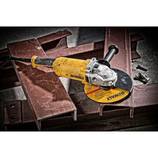 A DEWALT DWE490 angle grinder with a yellow and black design is placed on rust-colored metal beams, with some bolts nearby on a concrete floor. The grinder has a side handle and is shown in an industrial setting.