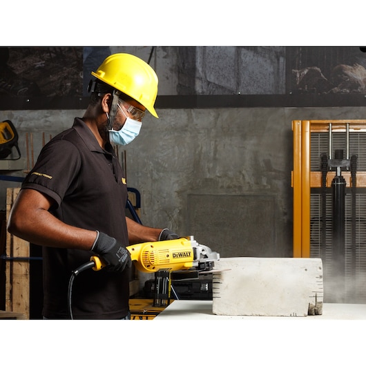 Worker uses DEWALT DWE492 tool to cut concrete block while wearing protective gear in an industrial setting.