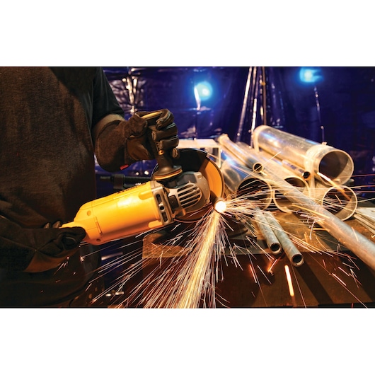 A person using a DEWALT angle grinder to cut metal pipes on a workbench, with bright sparks flying from the cutting area. Several metal pipes are stacked on the table in an industrial setting.
