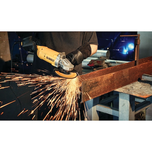 A person using a yellow DEWALT angle grinder to cut or grind a metal beam, with sparks flying from the contact point. The person is wearing protective gloves and working in a workshop setting.