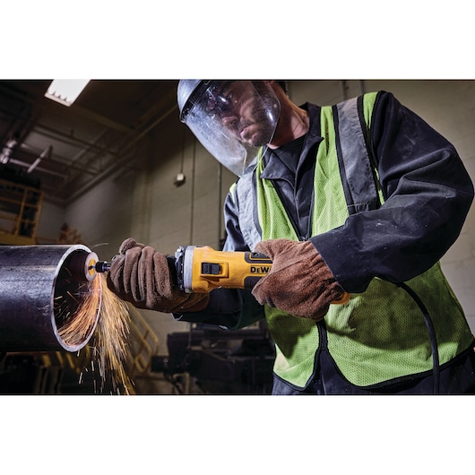 Worker using a DEWALT angle grinder to cut a metal pipe, sparks flying, wearing gloves and safety gear indoors.