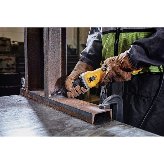 Person uses a DEWALT power tool to grind metal beams in an industrial workspace, wearing gloves and safety gear.