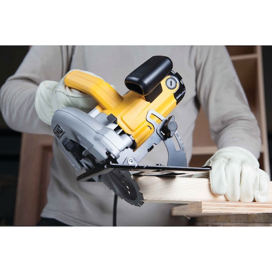 A person wearing gloves using a DEWALT DWE560 circular saw to cut a piece of wood. The saw is yellow and silver, and the person is working in a workshop setting.