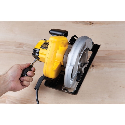 Hand uses screwdriver on a DEWALT DWE560 circular saw placed on a light wooden surface.
