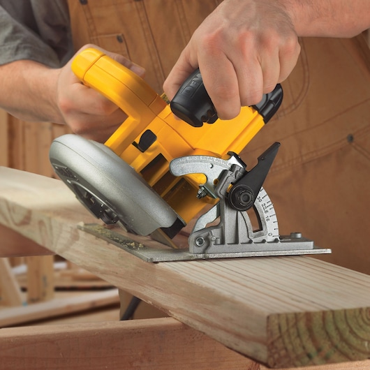 A person uses a yellow DEWALT circular saw to cut a wooden plank at an angle in a workshop setting.