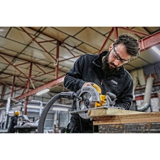 Worker using DEWALT DWE576K circular saw to cut wood in an industrial workshop, dust extraction hose attached.