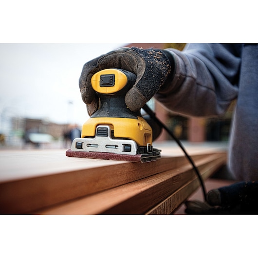 A close-up of a person wearing gloves using a yellow DEWALT electric sander to smooth the surface of a wooden plank.