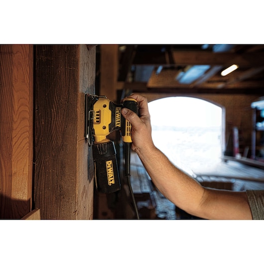 A person is using a DEWALT DWE6411 electric sander on a wooden surface inside a workshop, with the sander held vertically and dust bag attached.