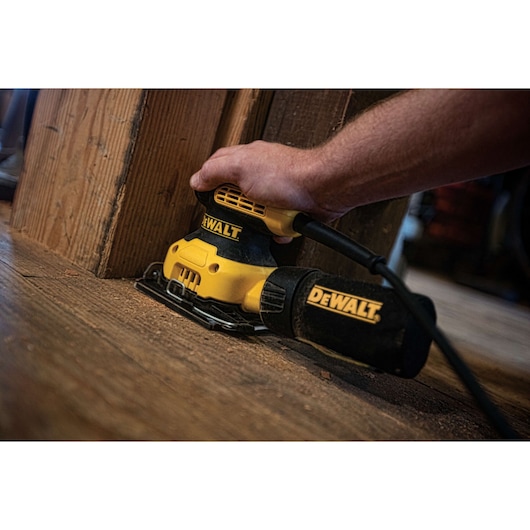 A close-up of a hand using a DEWALT electric sander on a wooden floor near a wooden post.