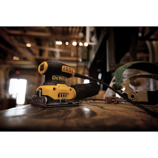 A DEWALT DWE6411 electric sander is placed on a dusty work surface in a workshop. The yellow and black tool is plugged in, with a safety mask and other equipment visible in the background.