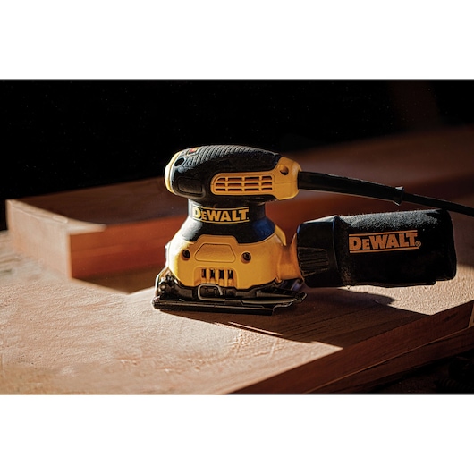 A DEWALT electric palm sander with a dust collection bag, placed on a wooden surface.