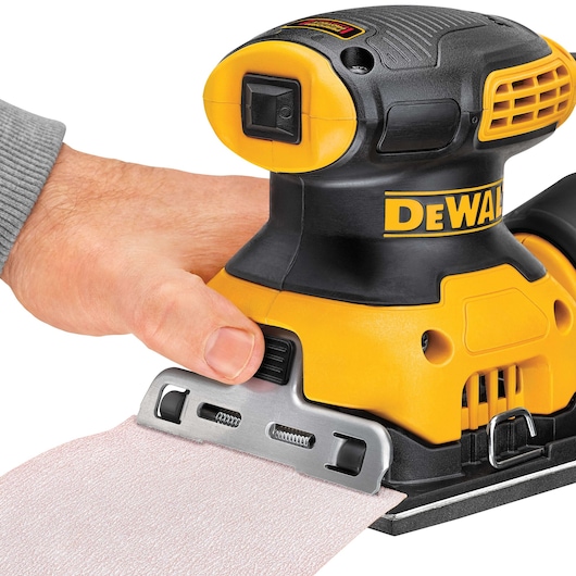 Close-up of a person's hand operating a DEWALT DWE6411 yellow and black electric sander, with a sanding sheet being attached to the tool.