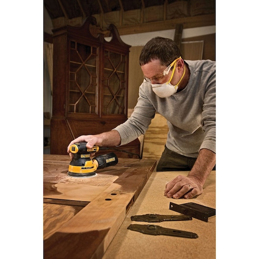 A person uses a DEWALT DWE6423 random orbital sander to smooth a large wooden surface in a workshop. The person’s face is blurred for privacy. Woodworking tools and cabinet parts are visible on the workspace.