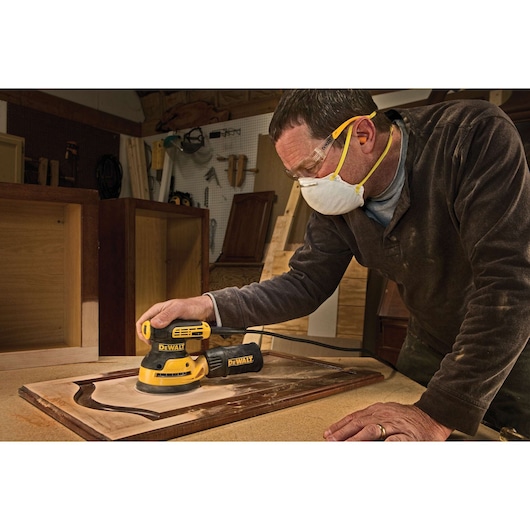 A person wearing a dust mask and safety goggles is using a DEWALT DWE6423 orbital sander to sand a wooden cabinet door in a workshop. The sander is yellow and black, and the environment includes woodworking tools and cabinets.