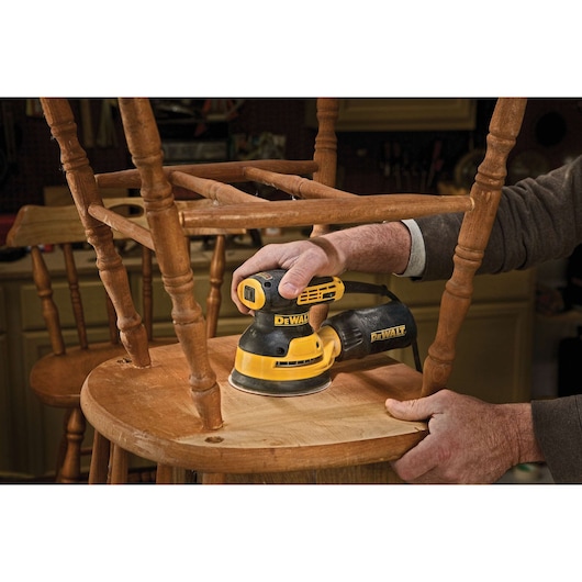 A person using a DEWALT DWE6423 electric sander to smooth the surface of a wooden chair in a workshop.