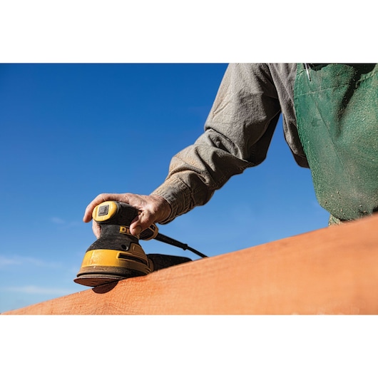 A person using a DEWALT electric sander on a wooden surface outdoors, under a clear blue sky. The person is wearing a green apron and a long-sleeve shirt.