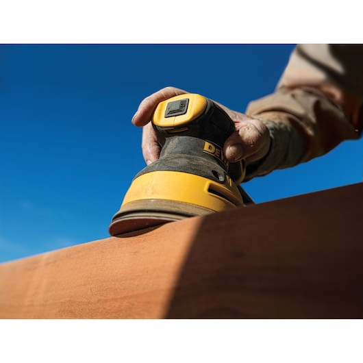 A close-up of a person using a DEWALT electric sander on a piece of wood, with a clear blue sky in the background.