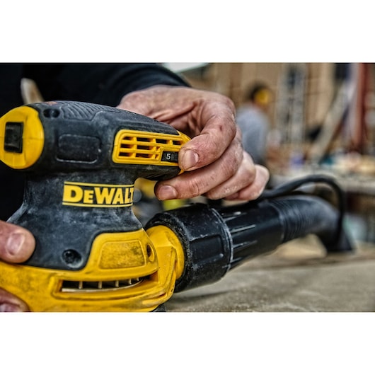 Hand adjusts DEWALT DWE6423 sander while working on a wood surface in a workshop.