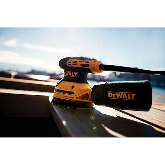 A DeWalt DWE6423 electric sander resting on a wooden surface outdoors, with sunlight illuminating the tool.