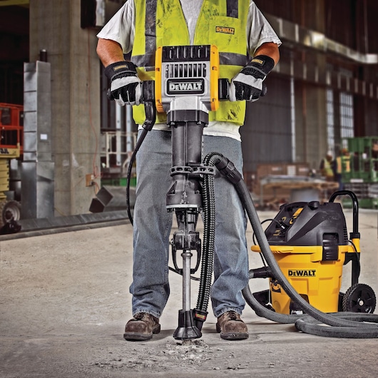 A person wearing construction gloves, boots, and a safety vest is operating a DEWALT demolition hammer on a concrete floor in an industrial workspace. Next to them is a DEWALT dust extractor vacuum connected to the tool by a hose.