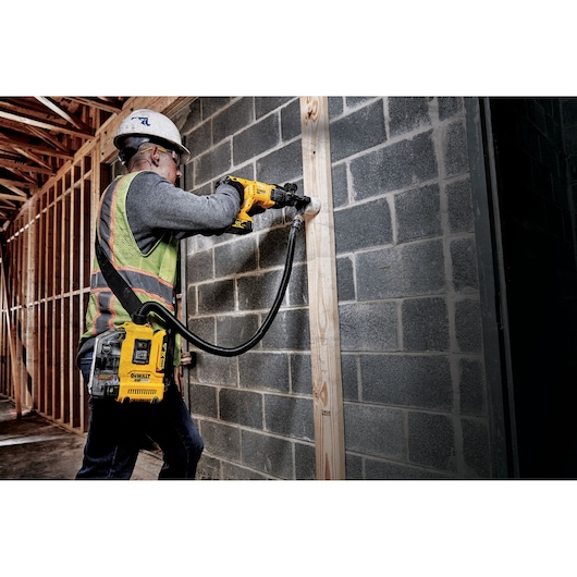 Worker drills into a concrete wall with a DEWALT tool and dust extractor in a construction site.