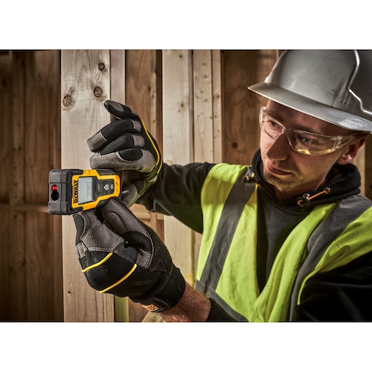 Worker uses DEWALT DWHT77100 laser distance measurer on wooden frame, wearing gloves and safety gear.