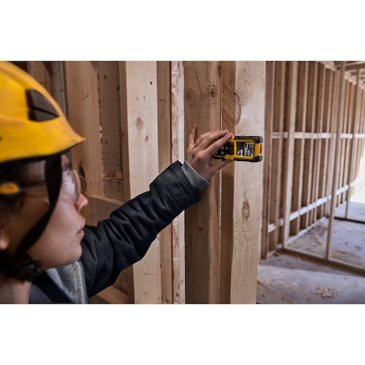 A person wearing a yellow hard hat uses a DEWALT DWHT78200 digital tool to measure or scan wooden framing at a construction site. The person's face is blurred for privacy.