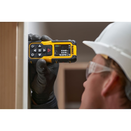 Gloved hand holds DEWALT laser distance measurer against wall; worker wears a white safety helmet.