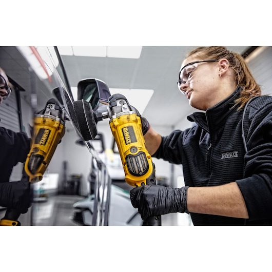 Person uses a yellow DEWALT polisher, model DWP849X, to polish a car side mirror indoors. Face is blurred.