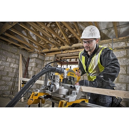 Worker using DEWALT DWS777 saw to cut wood at construction site. Safety gear and hi-vis vest worn.