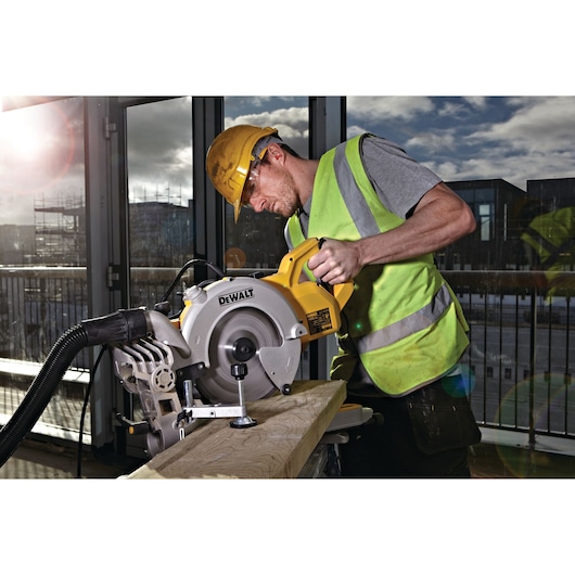 Worker in safety gear using a DEWALT DWS778 saw to cut wood indoors, with sunlight visible through windows.
