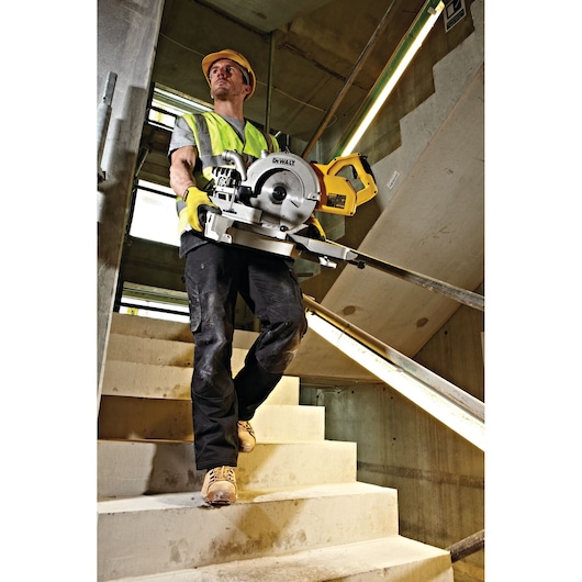 Worker in safety gear carries DEWALT DWS778 saw down concrete stairs at a construction site.