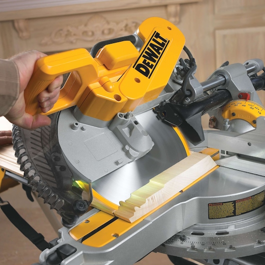 Close-up of a person using a yellow DEWALT miter saw (model DWS780) to cut a piece of wood. The saw is positioned on a workbench and the blade is in motion.