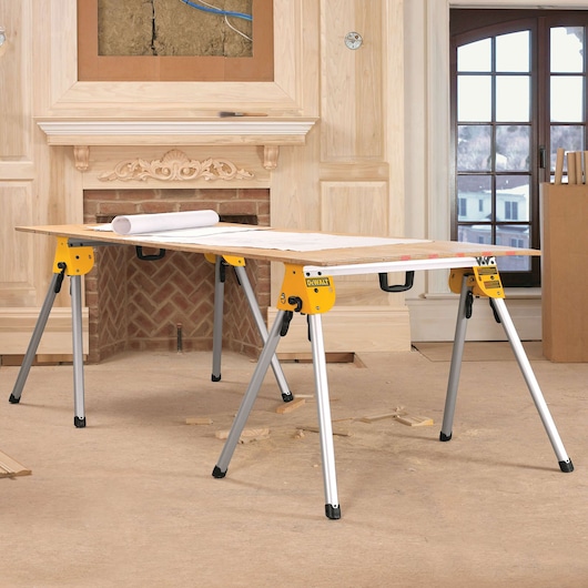 A DEWALT workbench with yellow metal sawhorse legs and a flat surface, shown inside a room under renovation. Blueprints and wood pieces are on the table, with construction materials visible in the background.