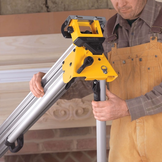 A person in work clothing is assembling a DEWALT miter saw stand, holding the yellow and black adjustable component with silver legs.