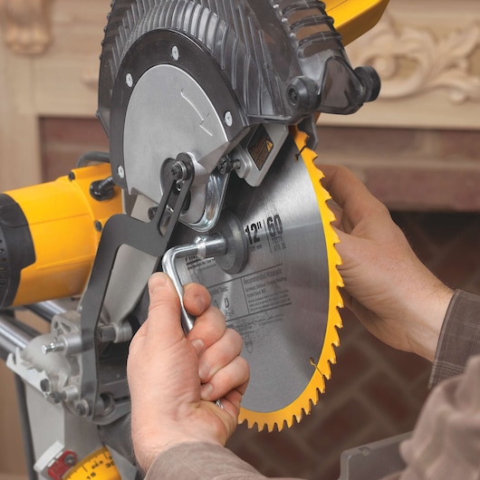 Close-up of hands changing the circular saw blade on a DEWALT DWS780 miter saw. The saw features a yellow casing and detailed mechanical parts.