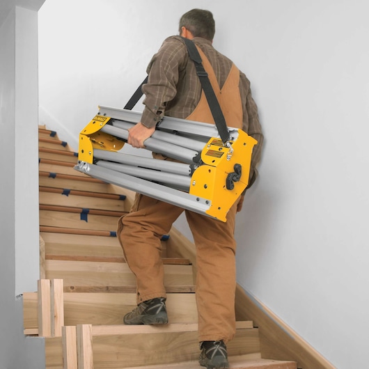 A person in work clothes carrying a folded DEWALT stand with a shoulder strap up a wooden staircase.