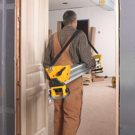 A person wearing brown overalls and a plaid shirt carries a DEWALT DWS780 tool stand with a carrying strap through a doorway in a construction or renovation setting.